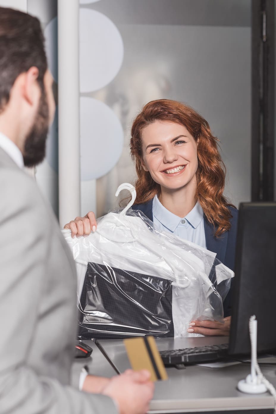 A saleswoman smiles while handing over freshly dry-cleaned clothes on hangers to a customer holding a credit card at a counter.