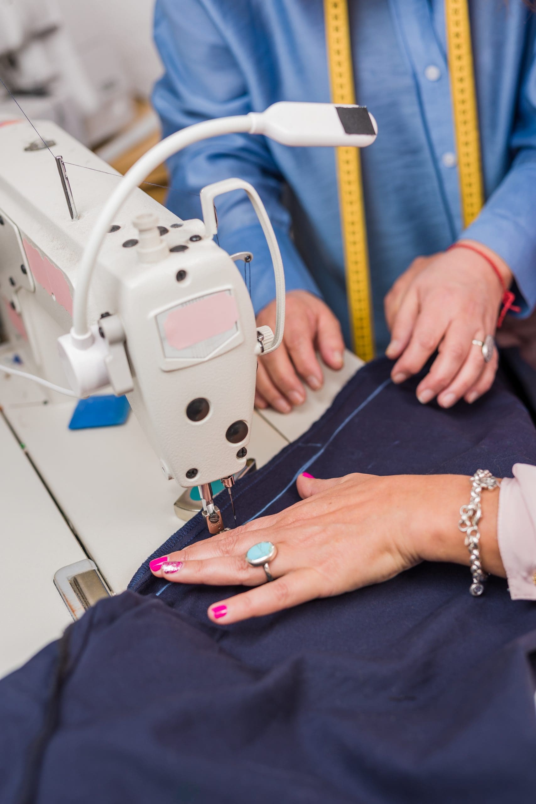 Two people use a sewing machine to stitch fabric; one person’s hands guide the material while the other supervises, and tools like a measuring tape are visible.