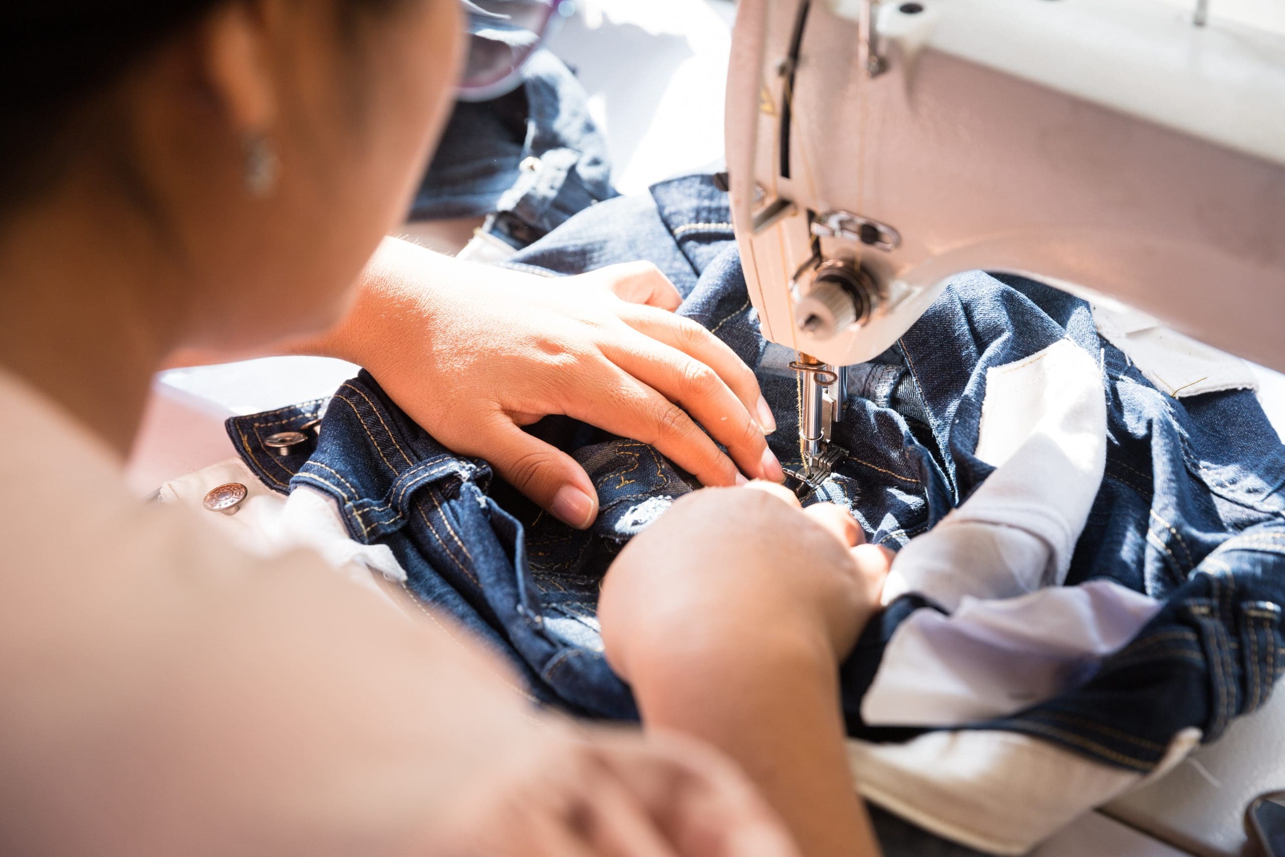 A person uses a sewing machine to stitch blue denim fabric, focusing on their hands guiding the material under the needle.