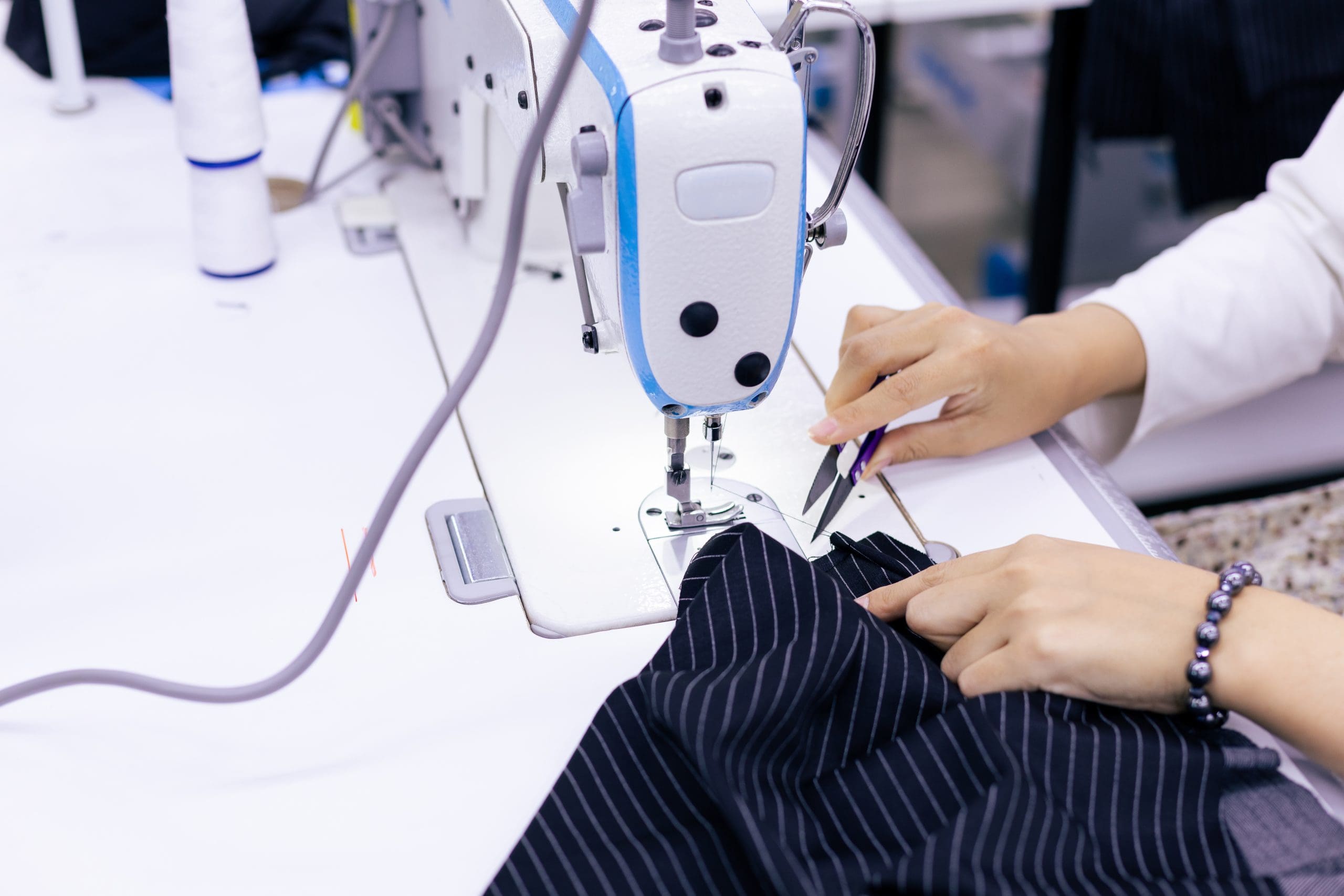 Person using a sewing machine to stitch black pinstripe fabric, with one hand guiding the material and the other holding small scissors.