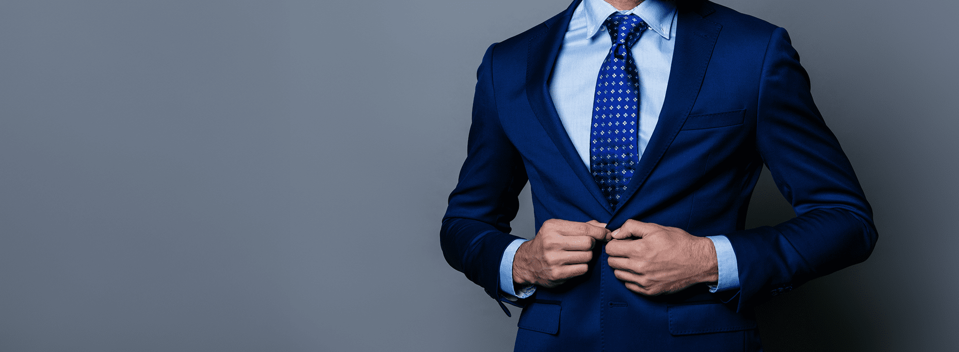Man in a blue suit and patterned tie buttoning his jacket against a plain gray background.