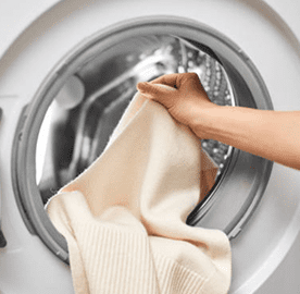 A person places a cream-colored sweater into the open door of a front-loading washing machine.
