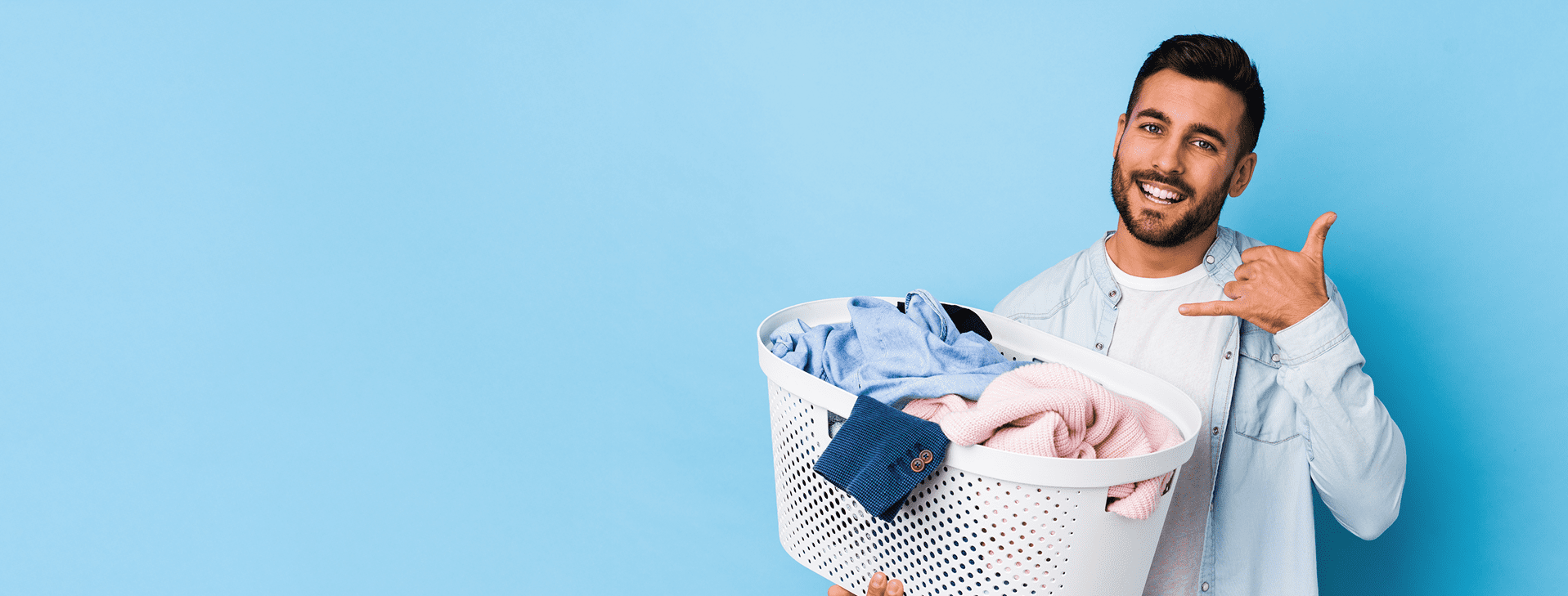 A man holds a laundry basket filled with clothes and makes a hand gesture resembling a phone call, standing against a blue background.