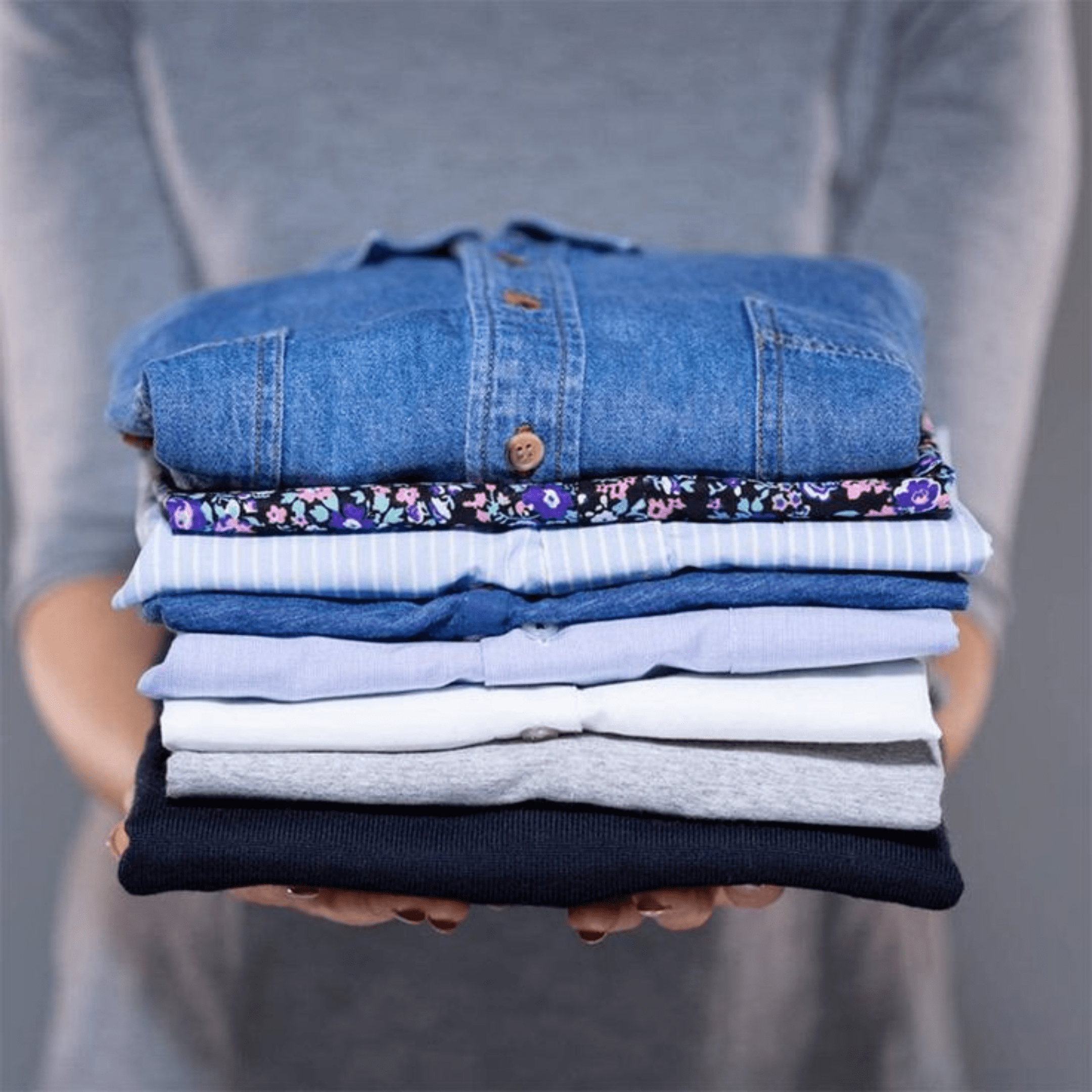 A person holding a neatly folded stack of assorted shirts and tops, including denim, floral, striped, and solid colors, against a plain background.