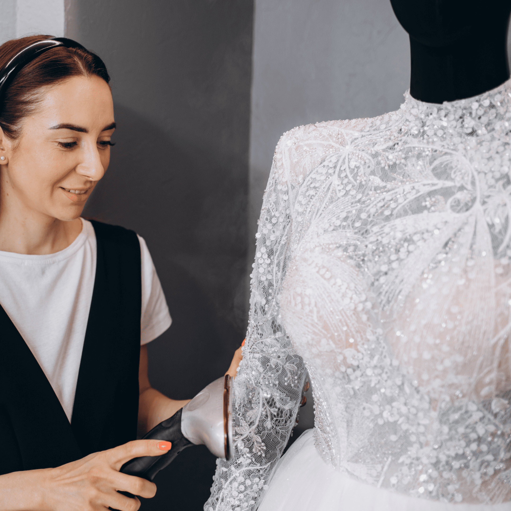 A woman uses a steamer to smooth the sleeve of a detailed, embellished white wedding dress displayed on a black mannequin.