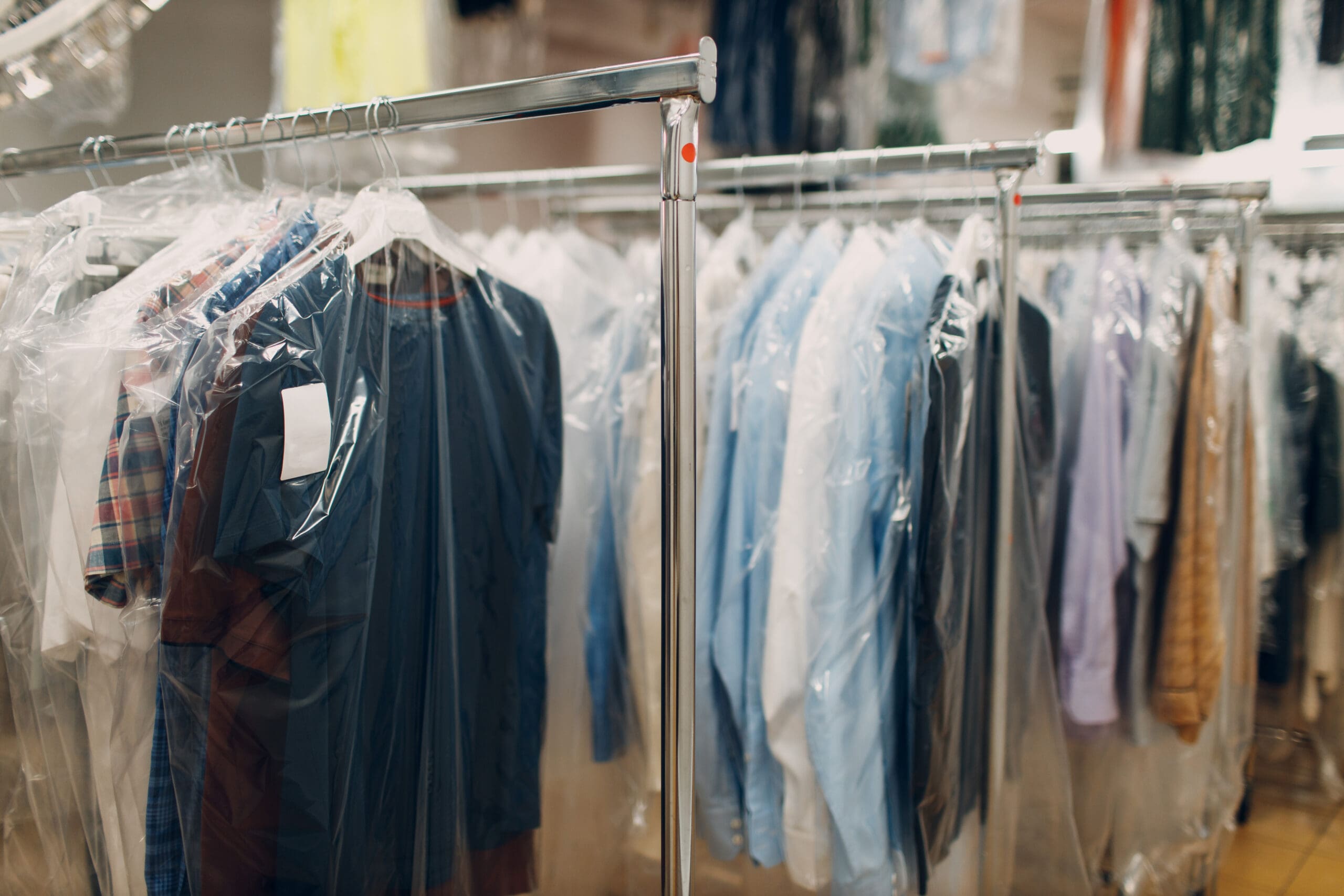 Clothes covered in plastic garment bags hang on metal racks inside a dry cleaning shop.