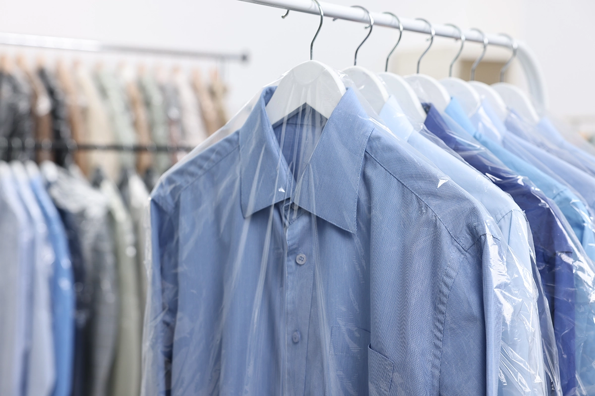 Light blue dress shirts on white hangers are covered with clear plastic garment bags, hanging on a clothing rack in what appears to be a dry cleaning or laundry facility.