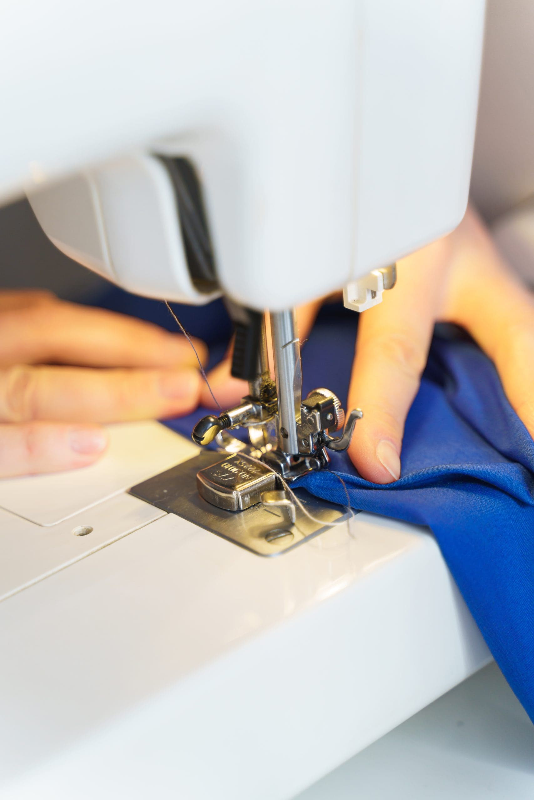 Close-up of hands guiding blue fabric through a sewing machine, with needle and presser foot in focus.