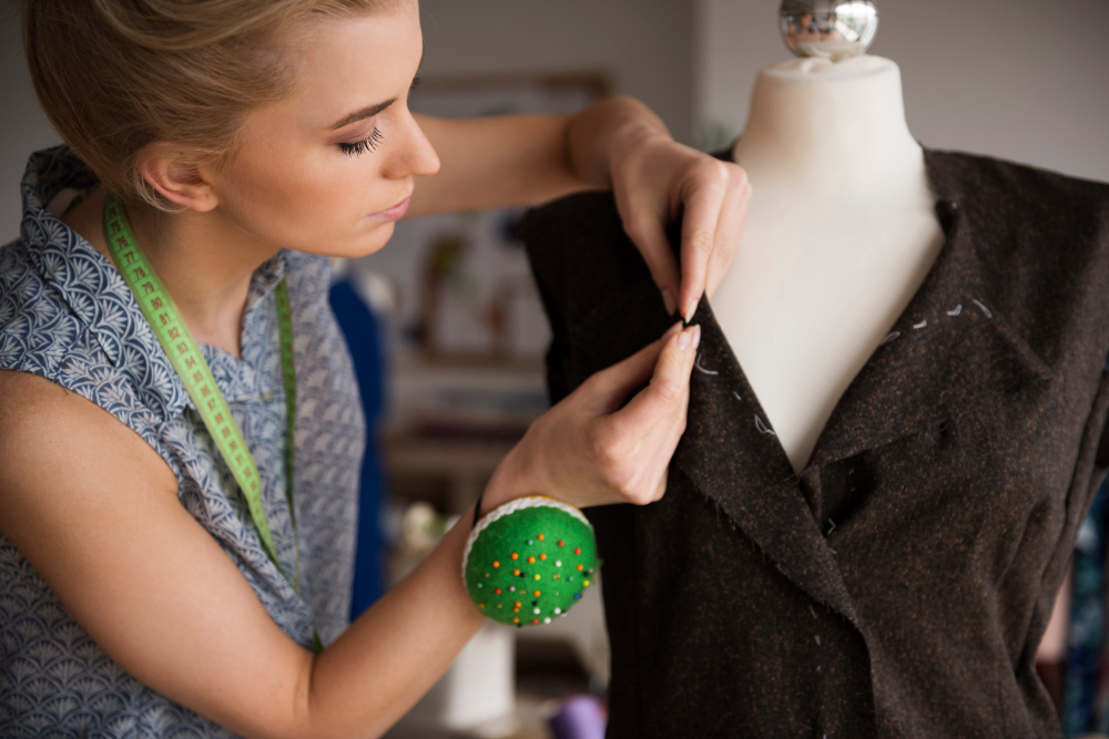 A person adjusts the lapel of a dark jacket on a dress form, wearing a measuring tape around their neck and a pincushion on their wrist.