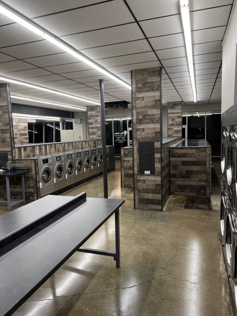 Modern laundromat interior with rows of washing machines, folding tables, and wood-paneled walls, brightly lit with overhead fluorescent lights.