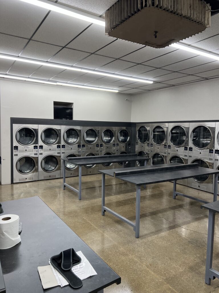 A clean, empty laundromat with rows of front-loading washers and dryers, folding tables in the center, and some supplies on a table in the foreground.
