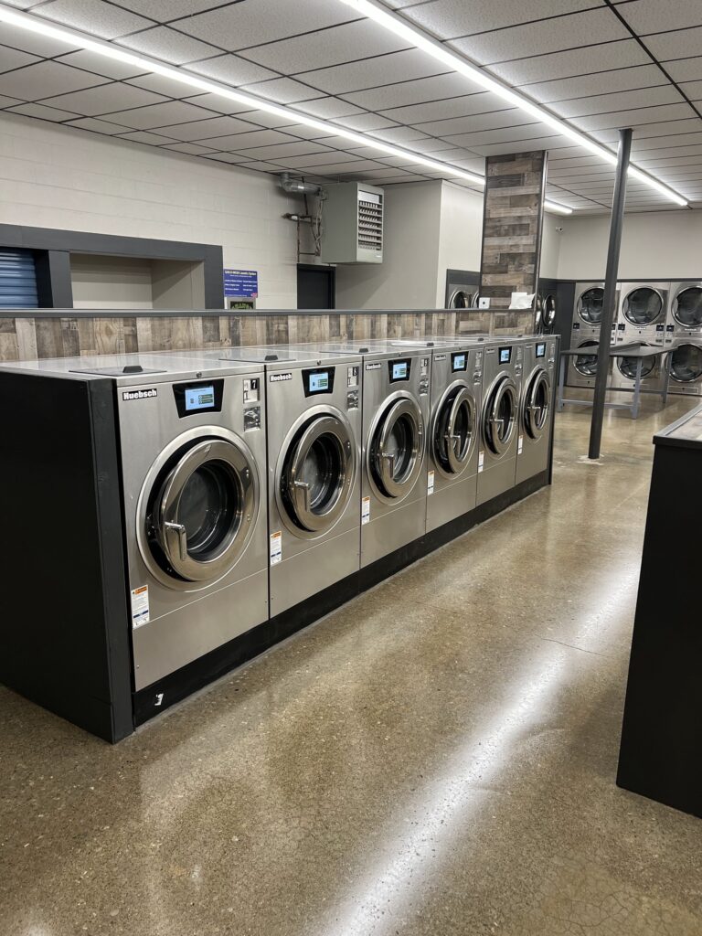 Row of six front-loading commercial washing machines in a clean, modern laundromat with polished floors and overhead fluorescent lighting.