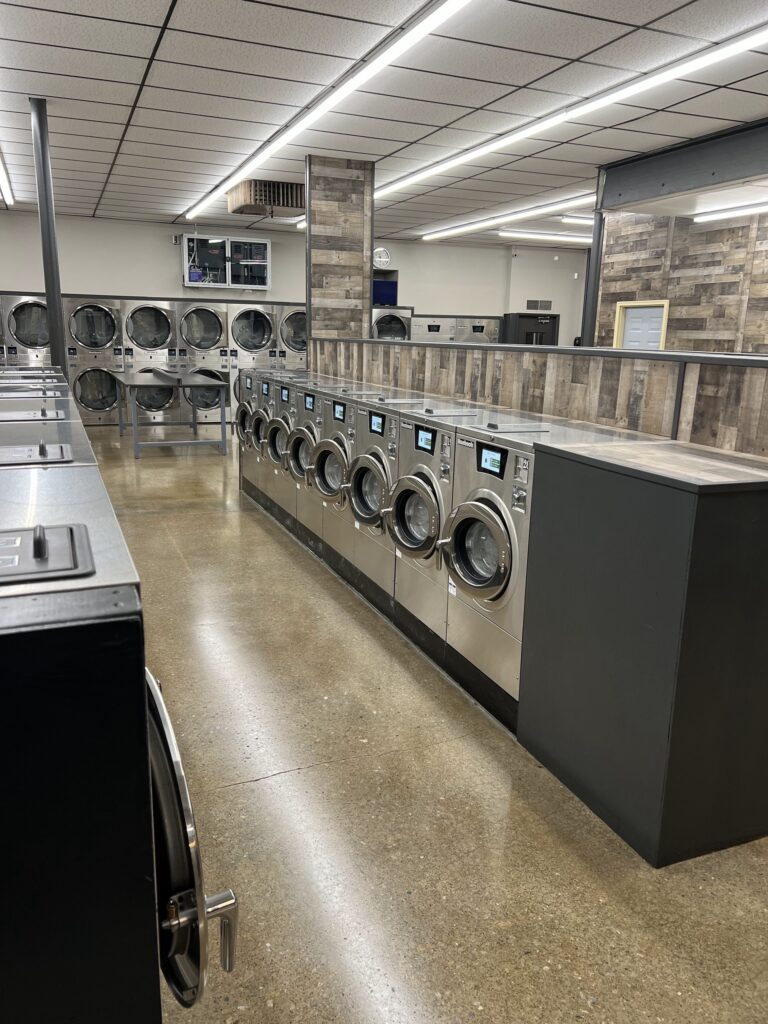 A row of front-loading washing machines and dryers in a clean, modern laundromat with polished floors and overhead lighting.
