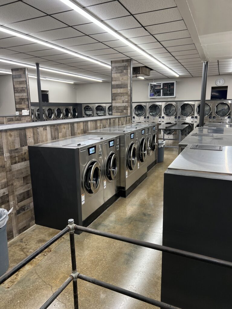 A clean laundromat with rows of stainless steel washing machines and dryers, polished concrete floors, and wood-accented walls.