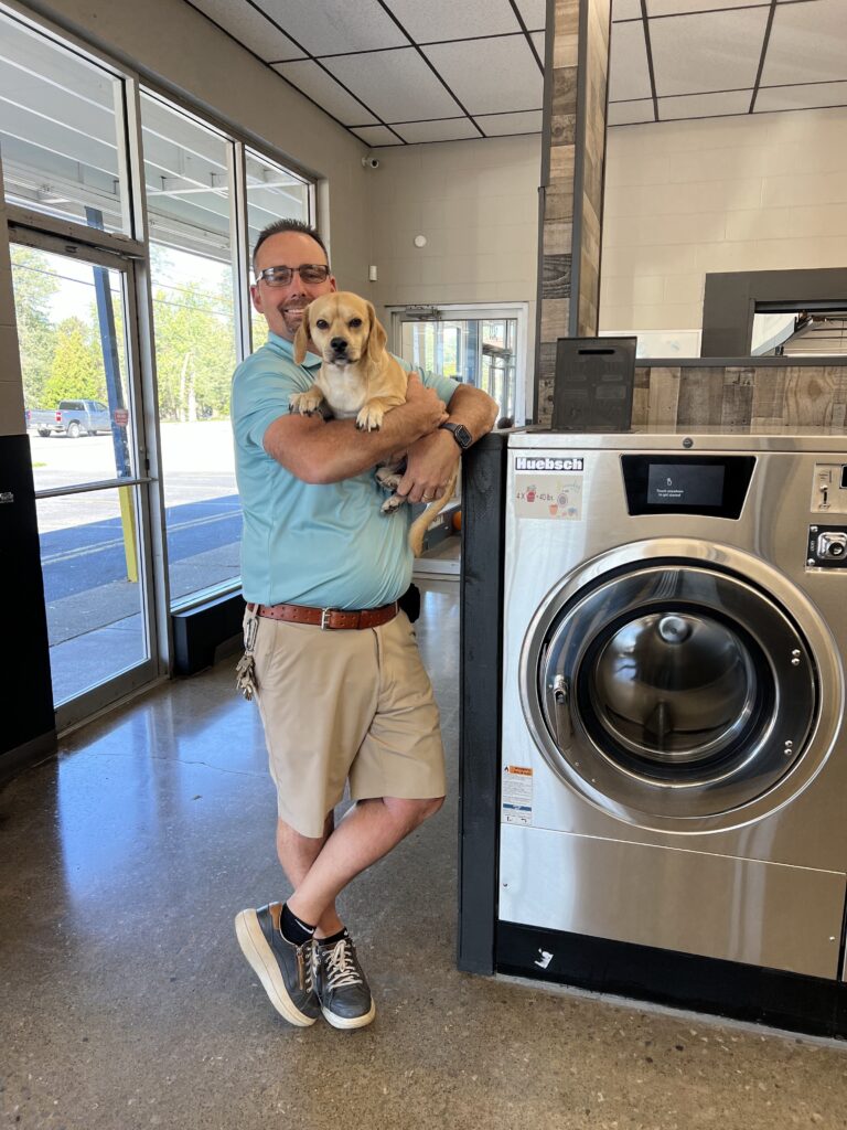 A man wearing glasses and a light blue shirt stands in a laundromat, smiling and holding a small tan dog next to a large washing machine.