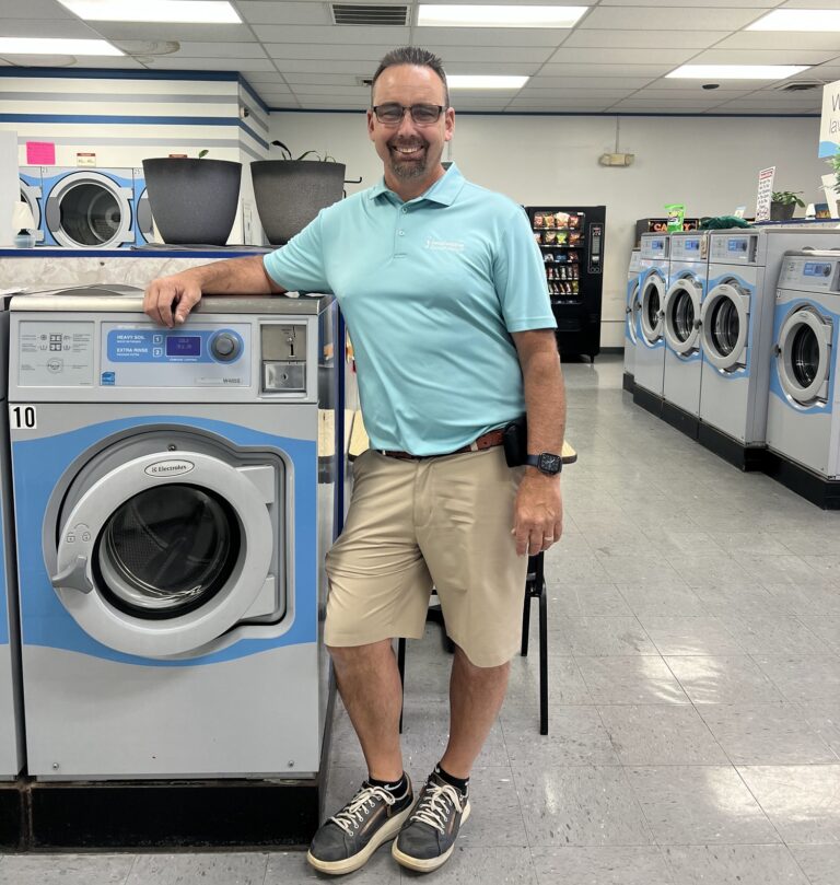 A man in a light blue polo shirt and khaki shorts stands smiling beside a front-loading washing machine in a laundromat.