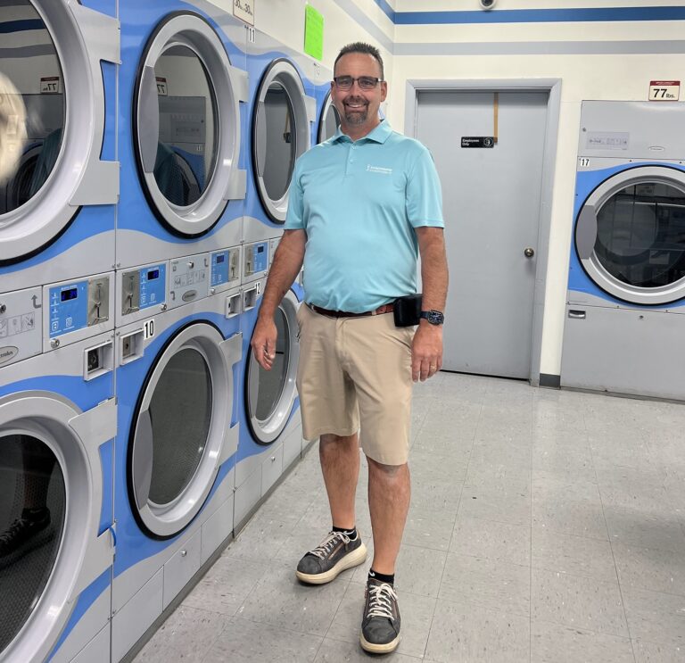 A man in a light blue polo shirt and khaki shorts stands beside a row of industrial dryers inside a laundromat.