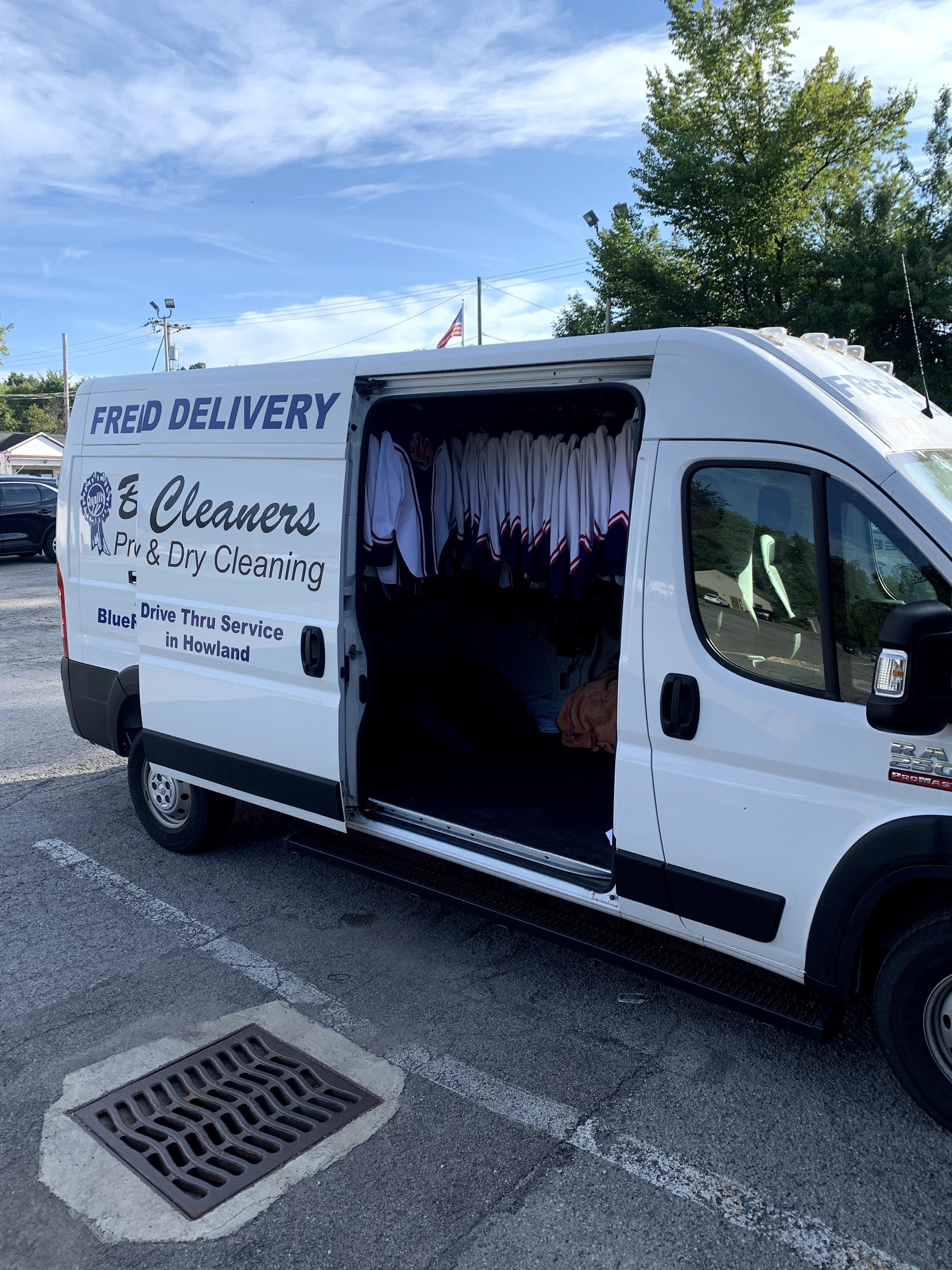 A white delivery van for "Freid Delivery & Cleaners" is parked with its side door open, revealing racks of hanging clothes inside.