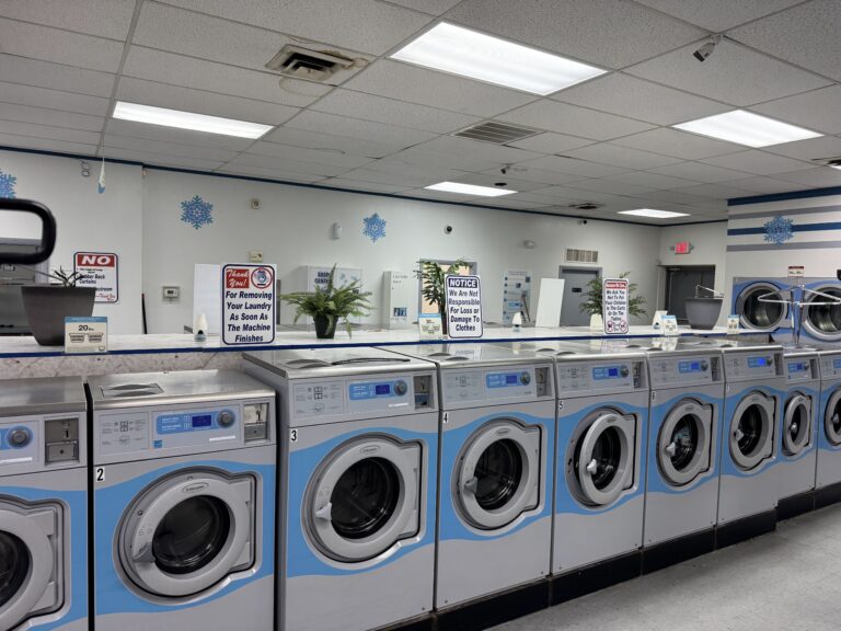 A row of front-load washing machines in a laundromat, with signs on top of the machines and a potted plant on the counter in the background.