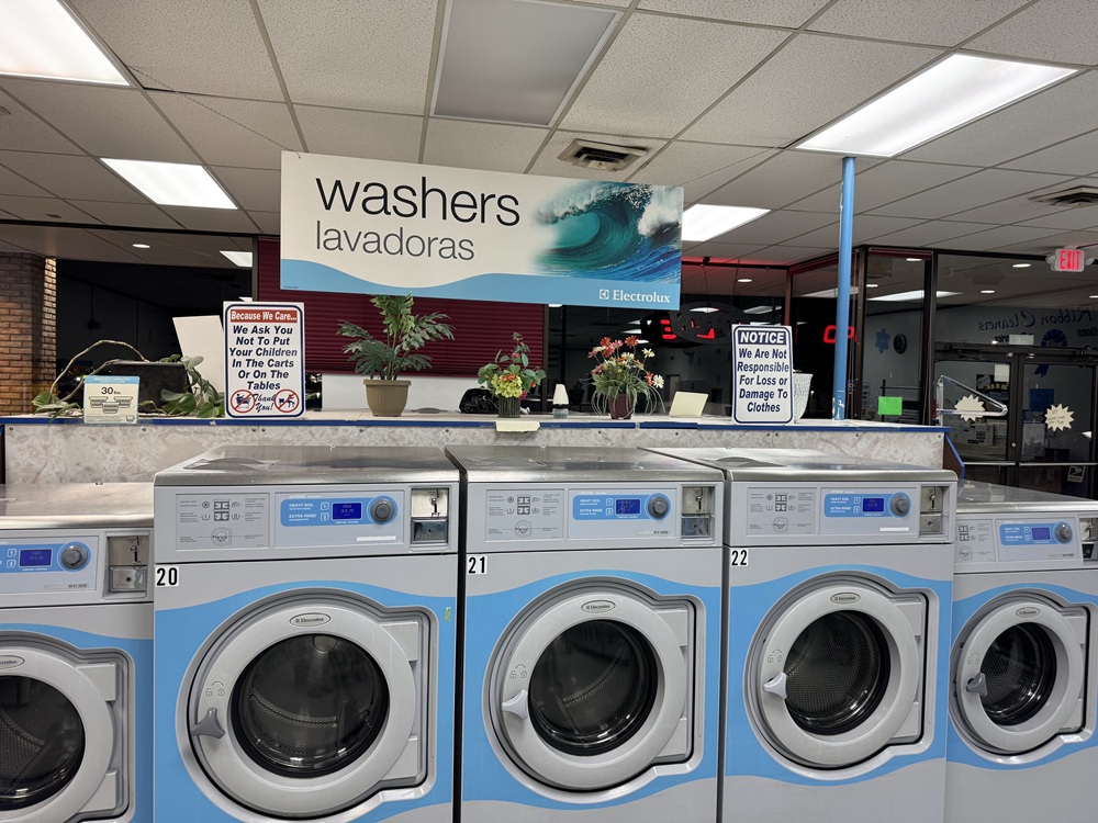 A row of front-loading washers numbered 20–22 in a laundromat, with a sign above reading "washers lavadoras" and several potted plants on a shelf.