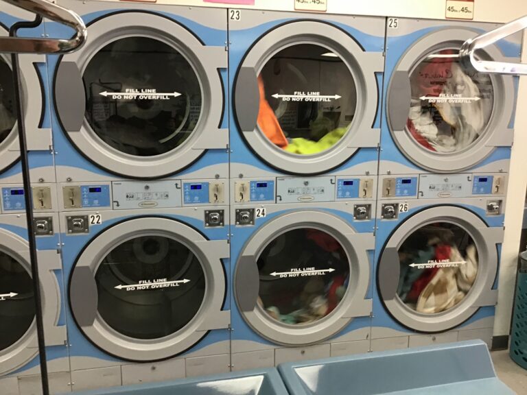 Six industrial front-loading dryers with laundry inside, stacked in two rows, with instructions printed on the doors and control panels above each machine.