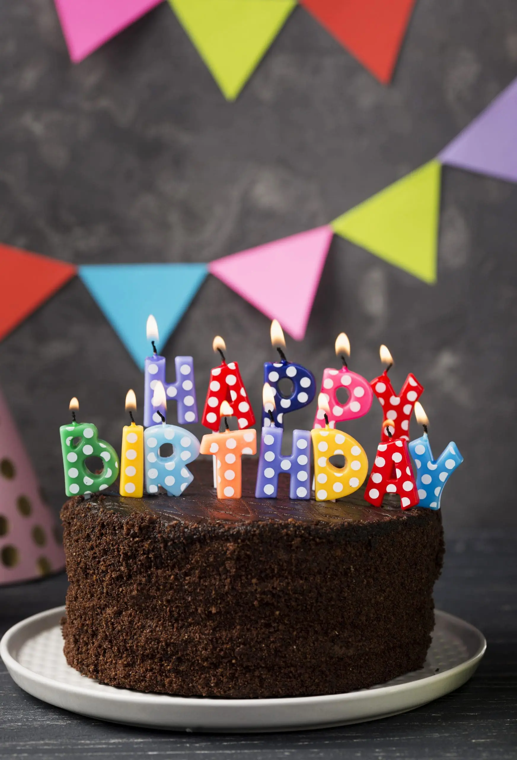 A chocolate cake with lit, colorful "HAPPY BIRTHDAY" candles on top, in front of a background with multicolored triangular banners.